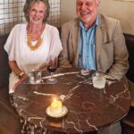A smiling woman in a white top and a smiling man in a light coloured suit and shirt, sitting at a restaurant table with drinks and a candle on