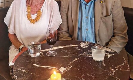 A smiling woman in a white top and a smiling man in a light coloured suit and shirt, sitting at a restaurant table with drinks and a candle on