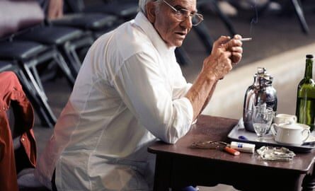Bradley Cooper in the film Maestro, sitting at a table and smoking a cigarette