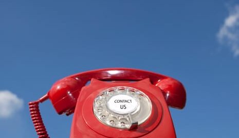 Red old-fashioned telephone with the words 'contact us' in the centre of the dialling pad, against a blue sky