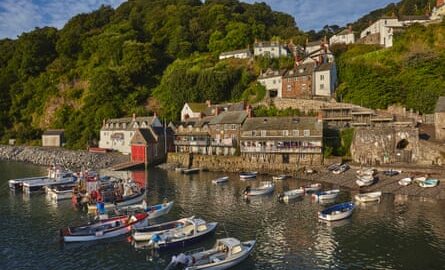 Boats moored in the sea in a stone harbour with old houses and cottages climbing the hillside beside them.