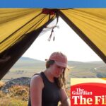Sian Lewis sits at a tent entrance using a camping stove, with hills in the background