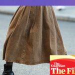 A person's legs in a long tan skirt and shiny black heeled ankle boots mid-stride