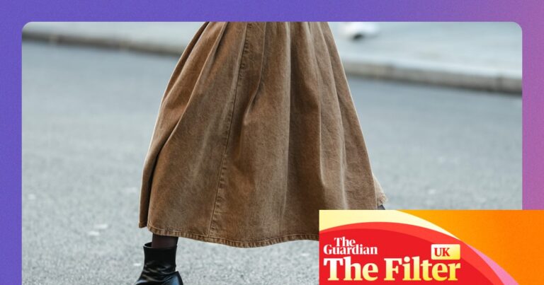 A person's legs in a long tan skirt and shiny black heeled ankle boots mid-stride