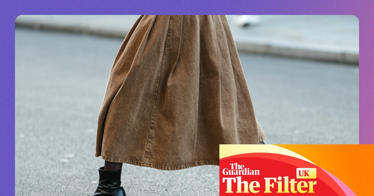 A person's legs in a long tan skirt and shiny black heeled ankle boots mid-stride