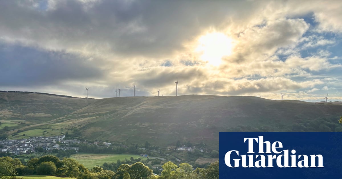 A view across a valley towards sunlit hills with wind turbines and a village off to the left